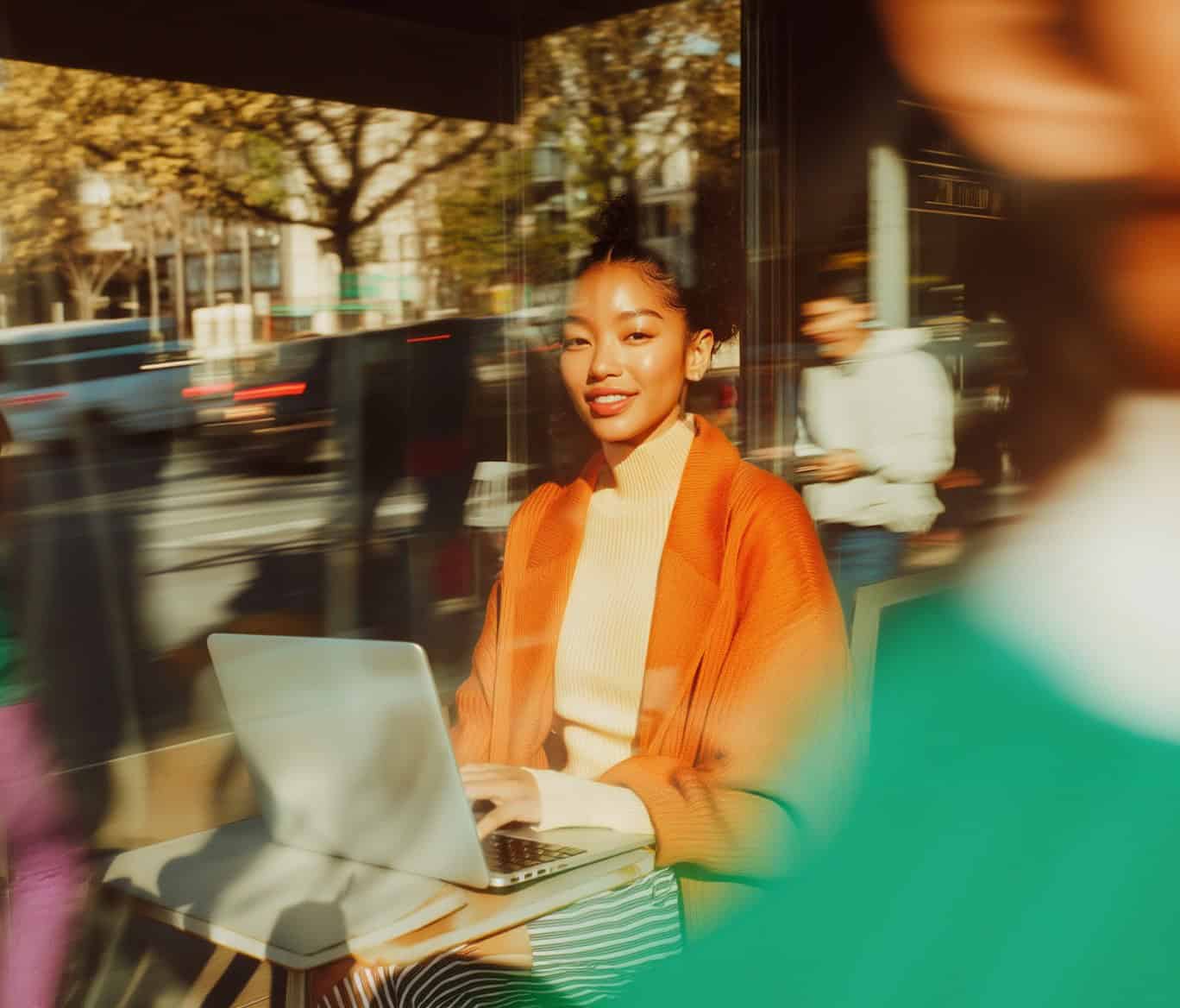 Young woman smiling while working on a laptop at a café, with city movement in the background.