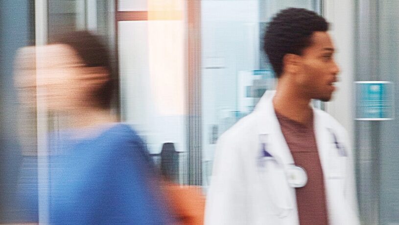 Two healthcare professionals walking through a modern hospital corridor, one wearing a white lab coat and a stethoscope, conveying a dynamic and technological environment.