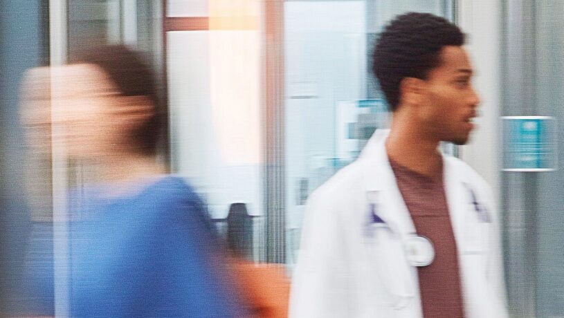 Dos profesionales de la salud caminando por un pasillo de hospital moderno, con uno de ellos llevando bata blanca y estetoscopio, en un entorno dinámico y tecnológico.